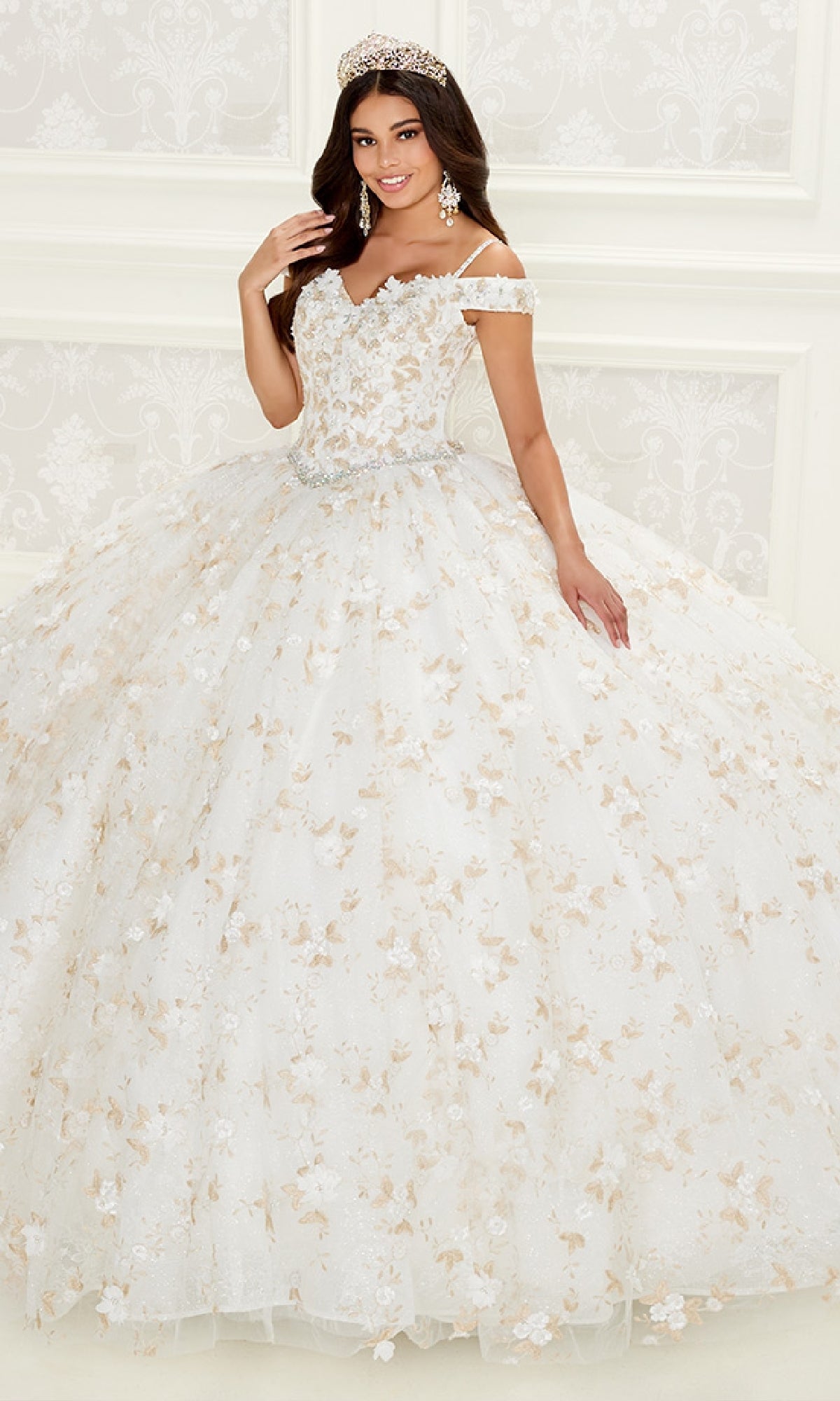 A woman in the PR30084 Quinceanera Dress by Princesa—an off-the-shoulder white gown with gold floral embroidery, corset back, and tiara—stands in a bright, ornate room.